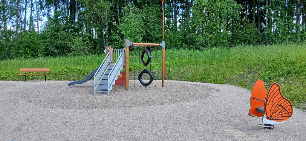Leikkipaikka, jossa on kiipeilyteline liukumäellä, rengaspuomeja, oranssi perhosaiheinen jousikeinu ja taustalla penkki sekä metsän reunaa.