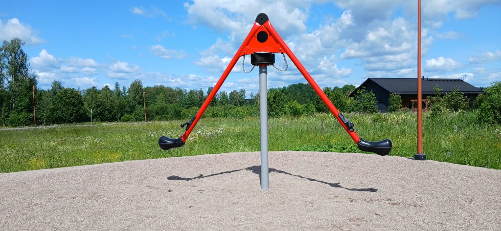 Uusi punamusta pyörähdyspeli hiekkapohjaisella leikkipaikalla kesäisessä maisemassa, taustalla niittyä, metsää ja tumma puutalo.