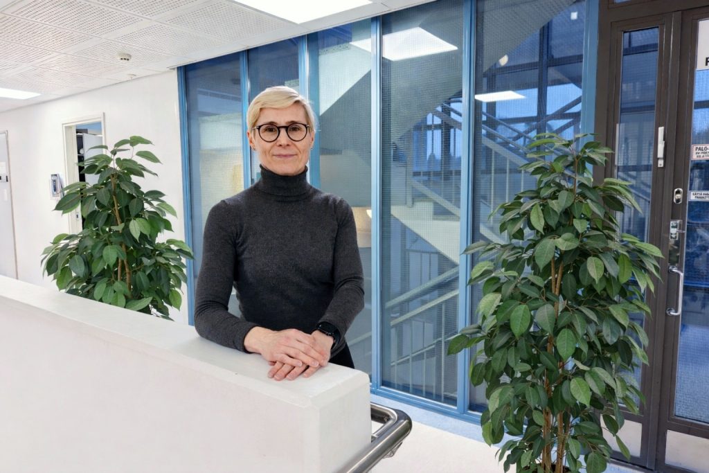 Lyhyttukkainen nainen seisoo modernissa toimistoympäristössä kaiteen äärellä. Taustalla on lasiseiniä ja viherkasveja.