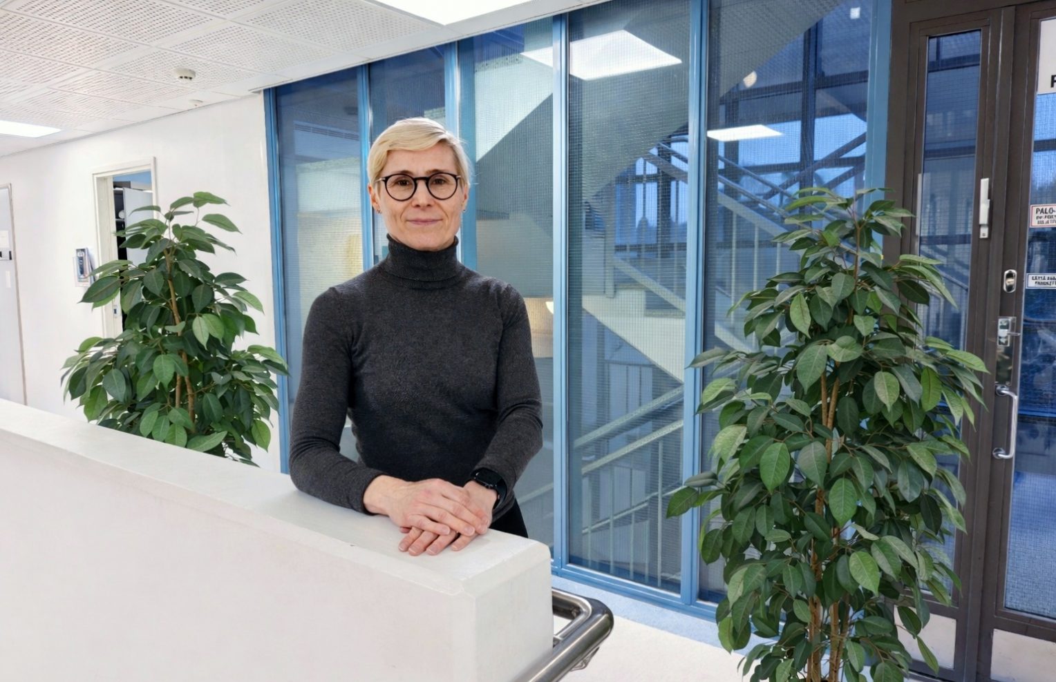 Lyhyttukkainen nainen seisoo modernissa toimistoympäristössä kaiteen äärellä. Taustalla on lasiseiniä ja viherkasveja.