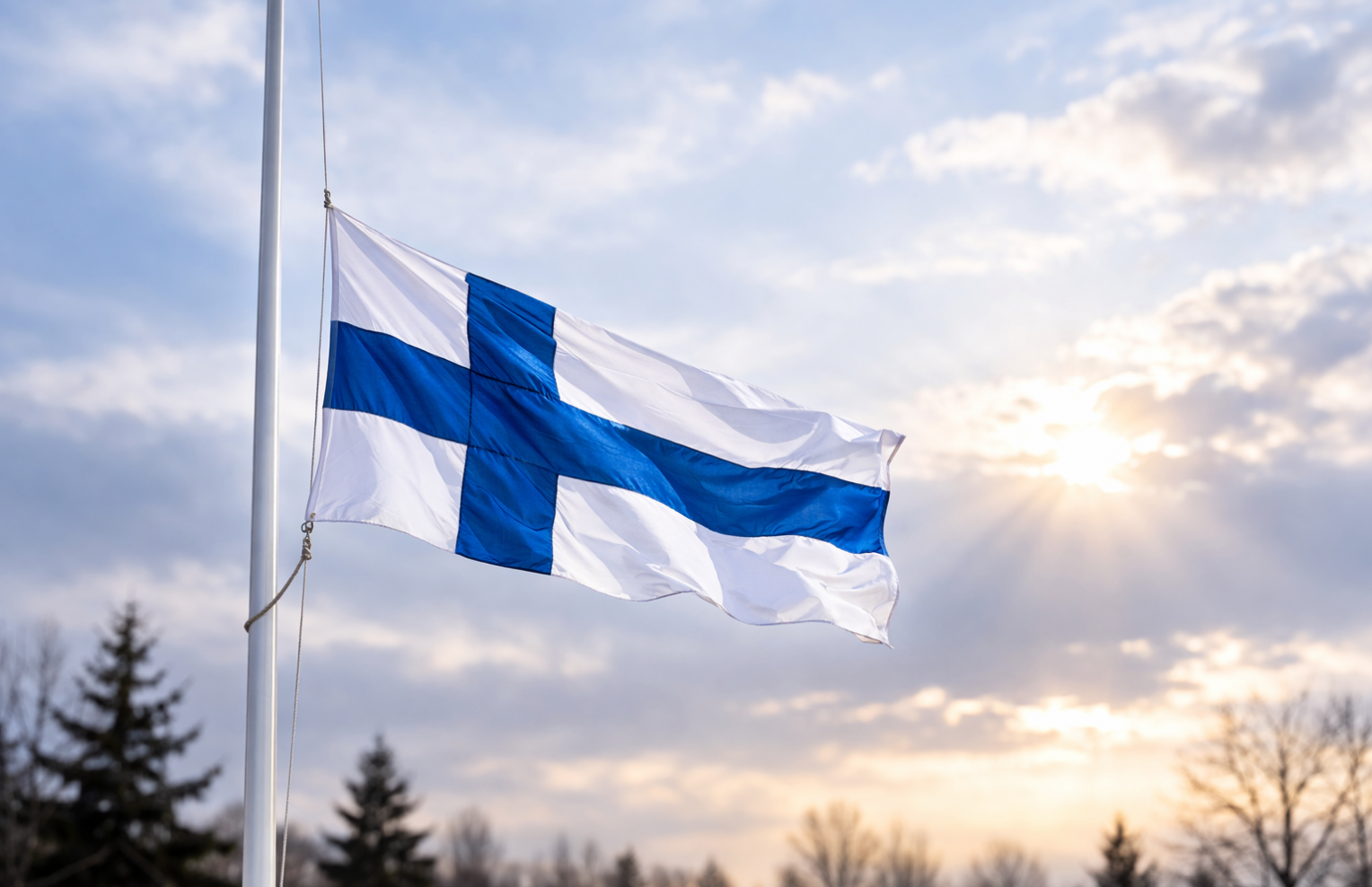 Suomen lippu puolitangossa lipputangossa, taustalla pilvinen taivas ja matalalta paistava aurinko.
