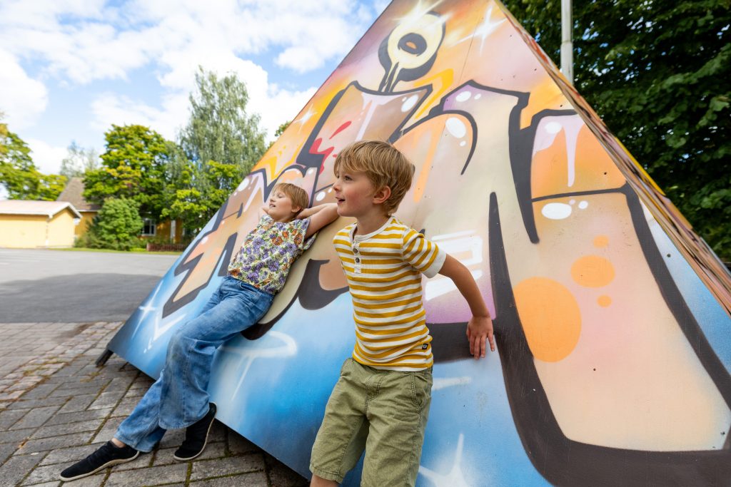 Kaksi lasta nojaa ulkona graffitein koristeltuun kolmion muotoiseen taideteokseen.