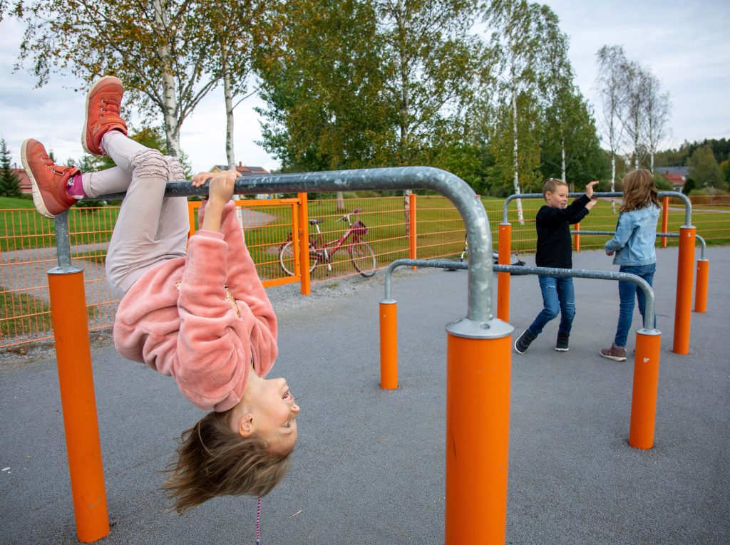Kolme lastaleikkipuistossa, yksi roikkuu pääalaspäin tangolla ja kaksi muuta leikkii taustalla.